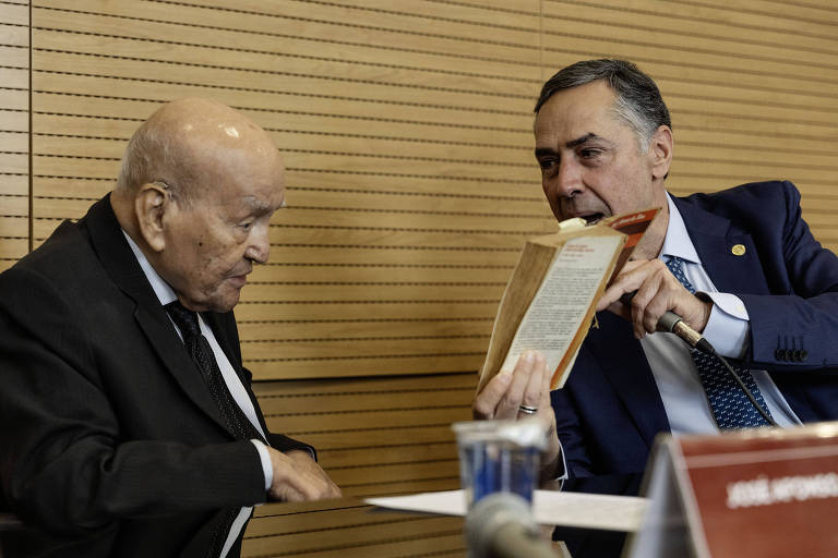 A imagem mostra dois homens sentados em uma mesa. O homem à esquerda, mais velho, está usando um terno escuro e parece estar ouvindo atentamente. O homem à direita, mais jovem, está vestido com um terno claro e está segurando um livro, mostrando-o para o homem mais velho. Ambos estão em um ambiente com paredes de madeira e uma mesa com copos de água.
