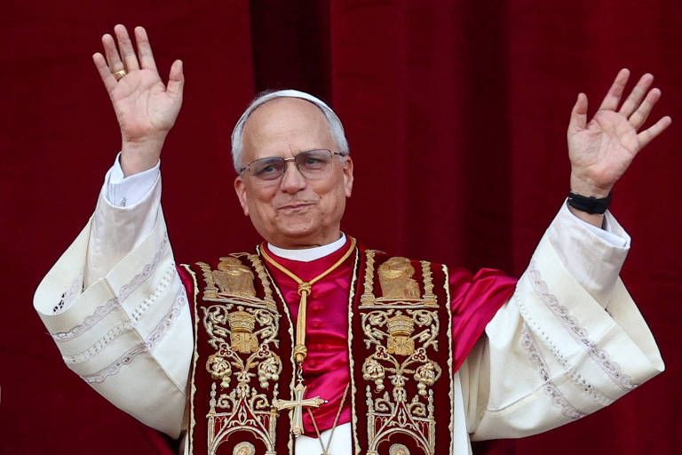 Um homem vestido com vestes religiosas, incluindo uma túnica branca e um manto vermelho, está acenando com as duas mãos levantadas. Ele usa óculos e tem cabelo grisalho. O fundo é de um tecido vermelho.