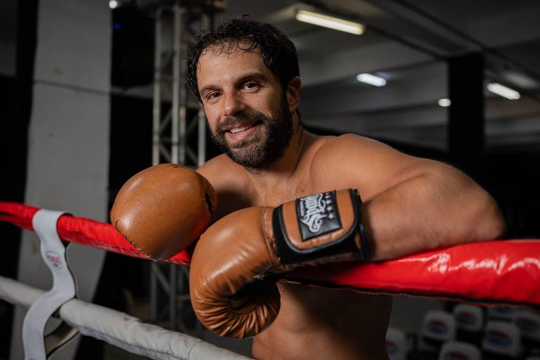 Um homem sorridente está posicionado em um ringue de boxe, apoiado nas cordas. Ele usa luvas de boxe marrons e está sem camisa, mostrando um físico atlético. O ambiente é bem iluminado, com estruturas metálicas ao fundo e assentos visíveis.
