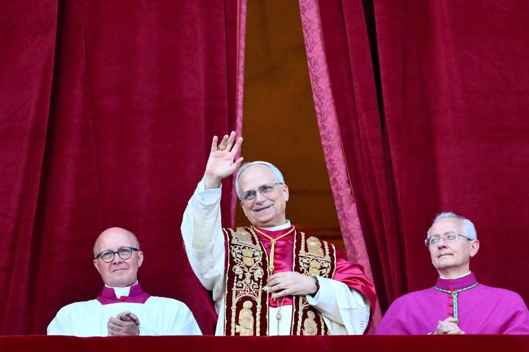 A imagem mostra um homem vestido com vestes papais, acenando com a mão direita. Ele está em uma varanda, cercado por duas outras pessoas, uma à sua esquerda e outra à sua direita, que também estão vestidas com roupas religiosas. O fundo é composto por cortinas vermelhas.