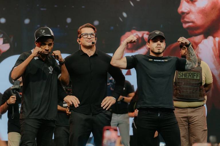A imagem mostra três homens em um evento de luta. Dois deles estão à esquerda e à direita, fazendo poses de força, enquanto o homem no meio, vestido com uma camisa preta, parece estar mediando ou apresentando. Ao fundo, há uma tela com uma imagem de um lutador. O ambiente é de um evento esportivo, com várias pessoas ao fundo, incluindo um segurança com um colete que diz 'SHERIFF'.