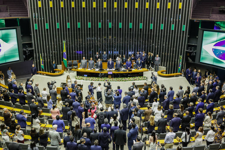 A imagem mostra uma sessão na Câmara dos Deputados do Brasil, com várias pessoas em pé e outras sentadas. No centro, há uma mesa com representantes, e ao fundo, bandeiras do Brasil e telas exibindo gráficos. A sala é ampla, com cadeiras dispostas em fileiras e um público que observa atentamente.