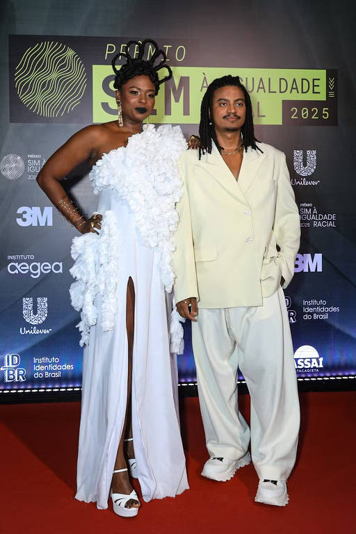 A imagem mostra duas pessoas em um evento de moda. A mulher à esquerda está vestindo um vestido branco com detalhes em flores e uma fenda na perna. Ela tem cabelo preso em um coque e usa brincos grandes. O homem à direita está usando um traje oversized em tom claro, com calças largas e uma camisa de botão. Ambos estão posando em um fundo com logotipos de patrocinadores e um tapete vermelho.