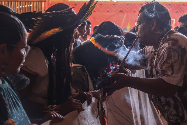 A imagem mostra um grupo de pessoas participando de uma cerimônia tradicional. Um homem está soprando fumaça de um instrumento, enquanto outras pessoas ao seu redor observam atentamente. Algumas pessoas usam trajes típicos e adornos, como penas. O ambiente parece ser um espaço fechado, com uma iluminação suave e um fundo desfocado.