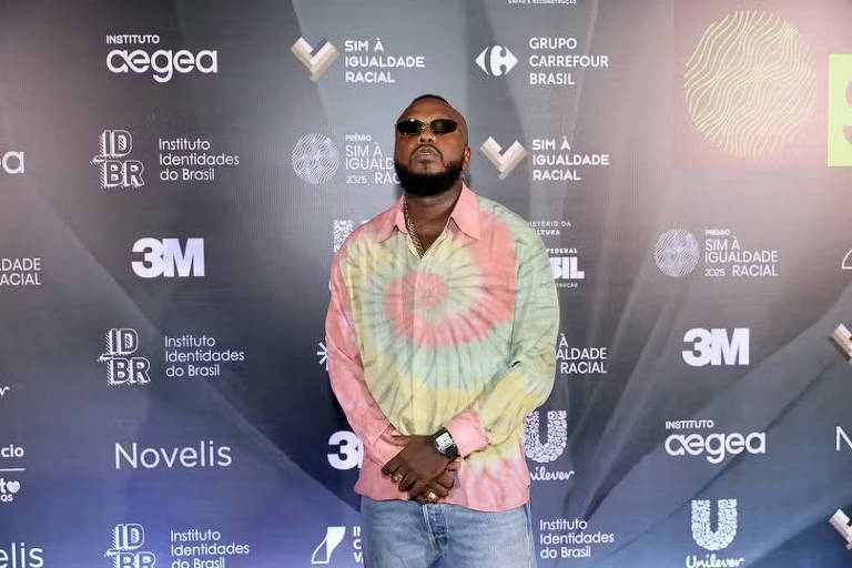 Um homem está posando em frente a um fundo com várias logomarcas, incluindo o Instituto Cegea, Instituto Identidades do Brasil, e Unilever. Ele usa uma camisa colorida com um padrão espiral e óculos escuros. O fundo é predominantemente cinza com elementos gráficos e textos relacionados à igualdade racial.