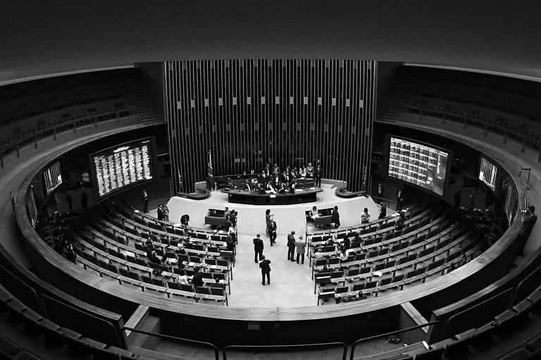 Imagem do plenário do Congresso Nacional do Brasil, com uma disposição circular. No centro, há uma mesa onde estão alguns representantes. As cadeiras estão dispostas em fileiras ao redor da mesa. Telas de exibição estão visíveis nas laterais, mostrando informações. O teto apresenta um design circular com um padrão em espiral.