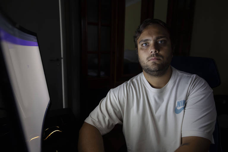 Um homem está sentado em frente a um computador em um ambiente escuro. Ele tem cabelo curto e uma barba bem aparada. O homem está usando uma camiseta branca com o logotipo da Nike em azul. A luz da tela do computador ilumina seu rosto, destacando sua expressão séria. Ao fundo, é possível ver uma porta e uma janela, mas estão em sombras devido à baixa iluminação.