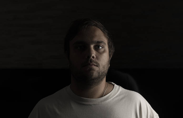 A imagem mostra um homem com cabelo curto e barba, olhando diretamente para a câmera. Ele está vestindo uma camiseta branca e o fundo é escuro, criando um contraste forte. A iluminação é suave, destacando seu rosto e expressões faciais, enquanto o restante da cena permanece em sombras.