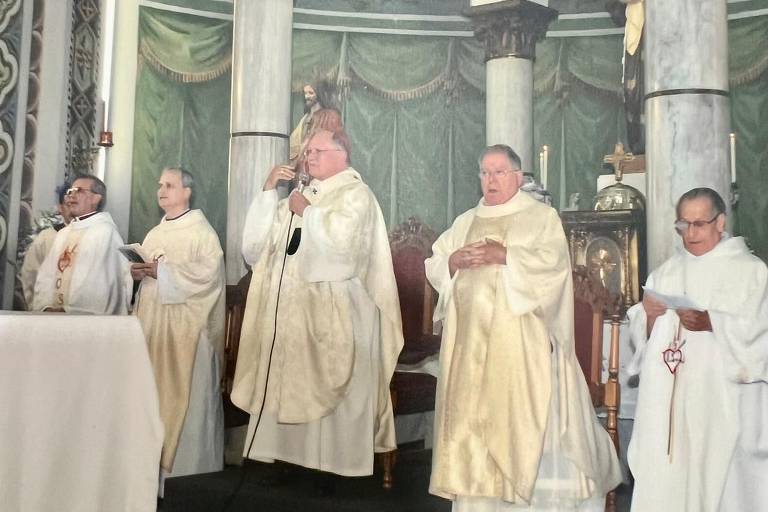A imagem mostra um grupo de cinco pessoas vestidas com vestes litúrgicas brancas, em um ambiente de cerimônia religiosa. Eles estão em pé em um altar, com um fundo decorado em verde e dourado. Um dos indivíduos está segurando um microfone e parece estar falando, enquanto os outros estão em posturas de escuta ou oração. Há também uma estátua visível ao fundo, além de velas e outros elementos decorativos no altar.