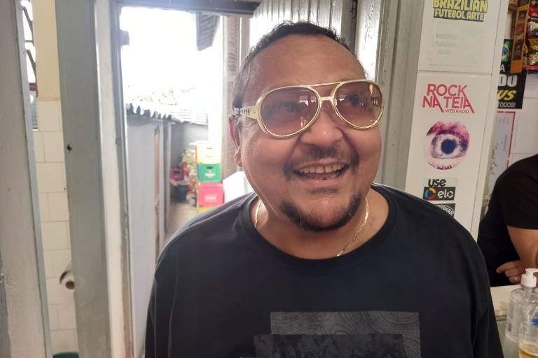 Um homem sorridente está posando para a foto, usando óculos grandes e redondos. Ele está vestindo uma camiseta preta e está em um ambiente interno, com uma porta ao fundo. Há alguns cartazes e adesivos nas paredes ao redor