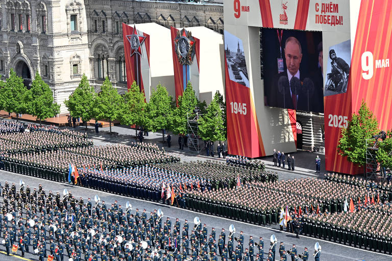 A imagem mostra um grande desfile militar em uma praça, com uma multidão de soldados em uniformes, organizados em fileiras. Ao fundo, há grandes telões exibindo uma mensagem em russo que diz 'Comemoração do Dia da Vitória!' e a data '9 de março'. Árvores estão alinhadas ao longo da praça, e edifícios históricos são visíveis ao fundo.