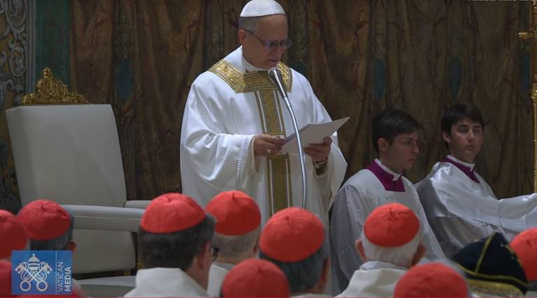 A imagem mostra uma cerimônia religiosa, onde um homem vestido com vestes litúrgicas brancas e douradas, que parece ser o Papa, está lendo um texto. Ele está em pé, segurando um papel, e é cercado por cardeais, que estão sentados em primeiro plano, usando capas vermelhas. Ao fundo, há uma decoração elaborada e uma cadeira alta ao lado do Papa. Dois assistentes estão próximos a ele, um deles vestindo uma túnica roxa.