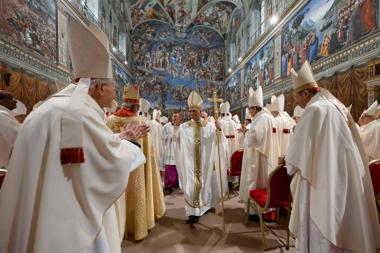 A imagem mostra uma cerimônia religiosa na Capela Sistina, com vários cardeais e bispos vestidos com vestes litúrgicas brancas e douradas. No centro, um sacerdote em vestes brancas está se movendo entre os outros clérigos, que estão aplaudindo. As paredes da capela estão decoradas com afrescos famosos, incluindo obras de Michelangelo. O ambiente é solene e reverente, com cadeiras vermelhas dispostas ao redor.