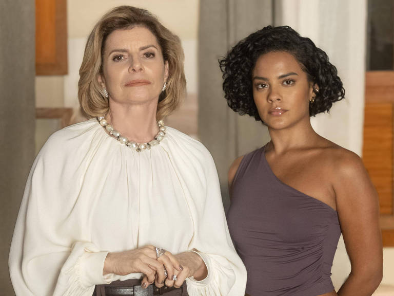 A imagem mostra duas mulheres em pé, uma ao lado da outra. A mulher à esquerda tem cabelo loiro e usa uma blusa branca com um colar de pérolas. A mulher à direita tem cabelo curto e cacheado, vestindo uma blusa de um ombro só na cor roxa. O fundo é composto por paredes claras e janelas de madeira.