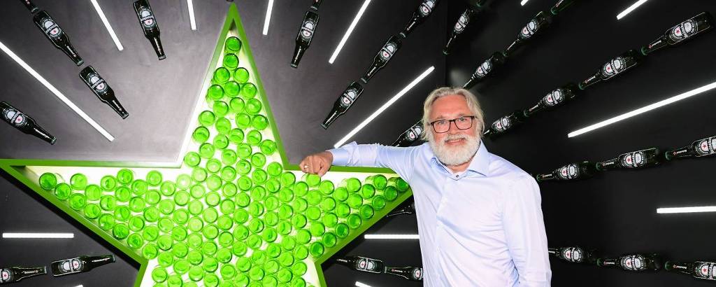 Um homem com cabelo grisalho e barba, usando uma camisa azul clara e calças escuras, está posando ao lado de uma grande estrela verde feita de copos ou objetos semelhantes. Ao fundo, há uma parede preta decorada com garrafas de cerveja dispostas em linhas verticais e luzes brancas que emitem um brilho. O homem está apoiando uma mão na estrela e sorrindo.