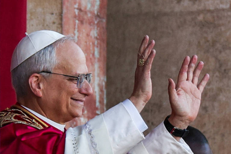 Um homem com uma vestimenta religiosa branca e vermelha, usando um chapéu branco, está acenando com as duas mãos. Ele tem óculos e um sorriso no rosto, em um ambiente que parece ser uma cerimônia religiosa. O fundo é de uma parede de pedra.