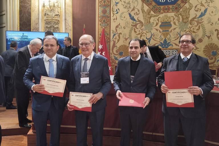 A imagem mostra quatro homens em um evento formal, todos segurando certificados. Eles estão em um ambiente decorado, com paredes ricamente adornadas e uma mesa ao fundo. Os homens estão vestidos com trajes formais, como ternos e gravatas. O primeiro homem à esquerda usa um terno azul, o segundo um terno escuro com gravata, o terceiro um terno preto e o quarto um terno escuro com colete. Todos parecem sorrir e estão em uma cerimônia de premiação ou reconhecimento.