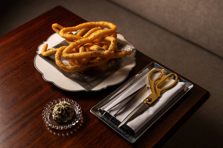 A imagem mostra uma mesa de madeira com um prato branco contendo um lanche em forma de espiral, que parece ser um tipo de biscoito ou salgadinho. Ao lado, há um pequeno prato com um creme ou pasta, e um conjunto de utensílios que inclui uma tesoura dourada, um garfo e uma faca, todos dispostos sobre um guardanapo branco.