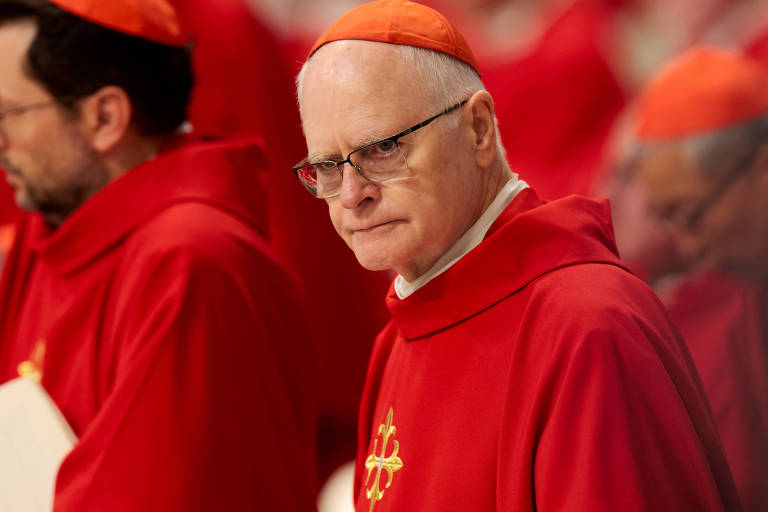 The image shows a group of cardinals dressed in red robes. One of the cardinals, in the foreground, is wearing glasses and has a serious expression. The background is blurred, but other cardinals can be seen also dressed in red. The atmosphere seems to be that of a religious ceremony.