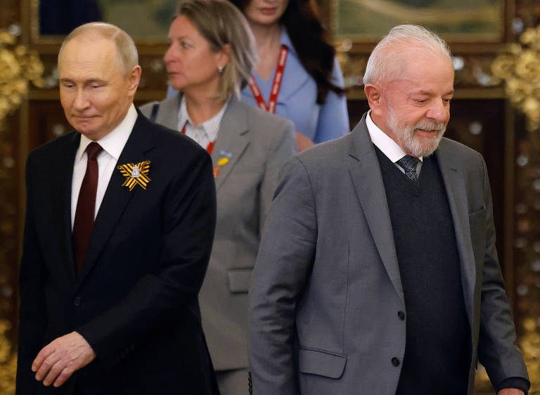 A imagem mostra dois homens caminhando lado a lado em um ambiente formal. O homem à esquerda, com um broche de fita, parece ser Vladimir Putin, enquanto o homem à direita, com um terno cinza, é Luiz Inácio Lula da Silva. Ao fundo, há uma mulher com cabelo liso e uma blusa azul, observando. O ambiente é decorado com elementos dourados.