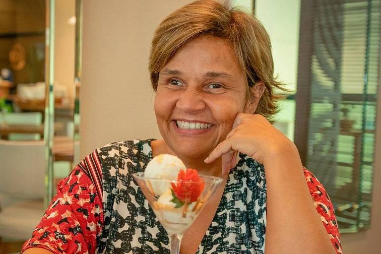 Uma mulher sorridente está sentada em uma mesa de restaurante. Ela tem cabelo curto e usa uma blusa com estampas coloridas. À sua frente, há uma taça de sobremesa com sorvete e uma fatia de morango. A mesa é de madeira e o ambiente parece ser bem iluminado e moderno.