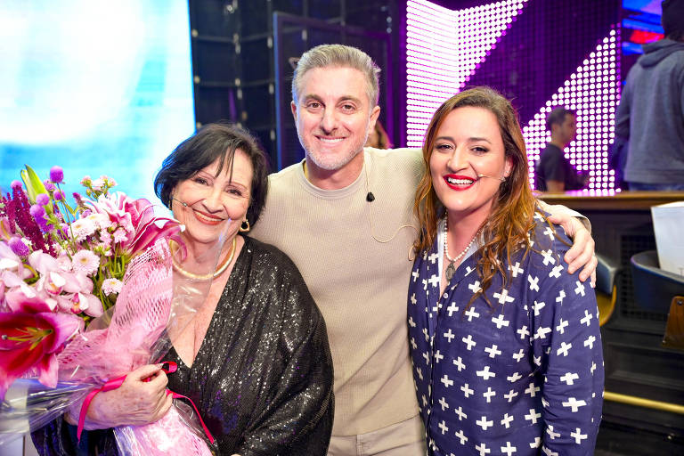 Três pessoas posam para a foto em um ambiente de evento. A mulher à esquerda segura um buquê de flores e usa uma blusa preta brilhante. O homem no centro veste uma roupa clara e tem um sorriso. A mulher à direita usa um pijama azul com estampas brancas e também sorri. O fundo é iluminado com luzes coloridas.