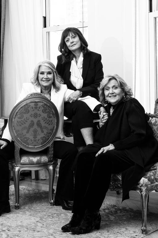A imagem em preto e branco mostra três mulheres em um ambiente interno elegante. Duas delas estão sentadas, uma à esquerda e outra à direita, enquanto a terceira está em pé, apoiada em uma cadeira. As mulheres estão vestidas de forma sofisticada, com roupas escuras e claras. O fundo apresenta cortinas e uma janela, sugerindo um ambiente bem iluminado. O piso é coberto por um tapete claro.