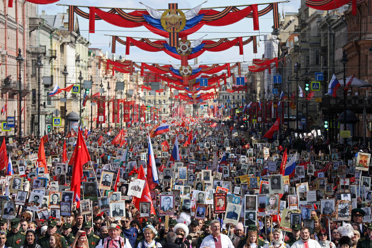 Uma grande multidão participa de uma manifestação em uma rua decorada com bandeiras e faixas. As pessoas seguram retratos de indivíduos, possivelmente de familiares ou figuras históricas, enquanto marcham. O ambiente é festivo, com muitas bandeiras vermelhas e azuis visíveis ao longo da rua, e o céu está claro.