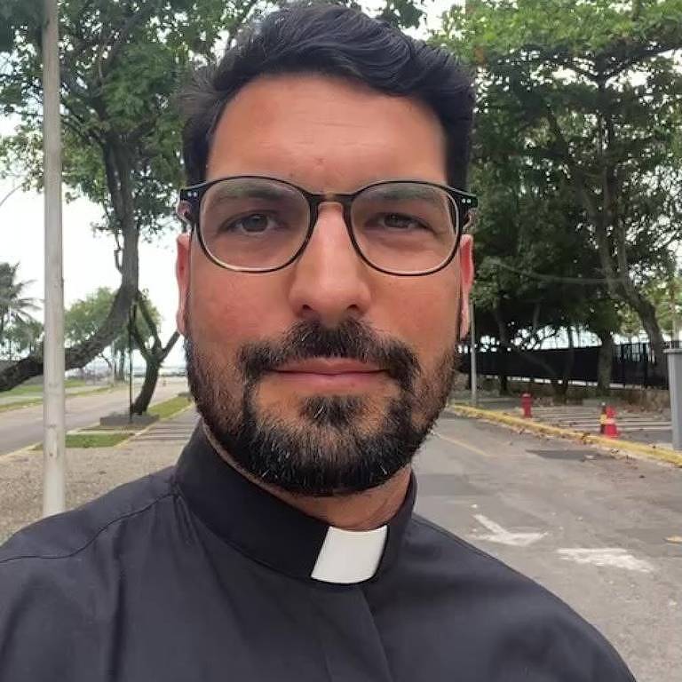 Um homem com barba e óculos, vestindo uma camisa preta com colarinho clerical branco, está posando para a foto. Ao fundo, há árvores e uma rua pavimentada, sugerindo um ambiente ao ar livre.
