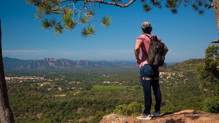 Um homem está em pé em uma rocha, olhando para uma vasta paisagem verdejante com montanhas ao fundo. Ele usa uma camiseta rosa e uma mochila nas costas. A cena é iluminada por um céu claro e azul, com algumas árvores ao redor.