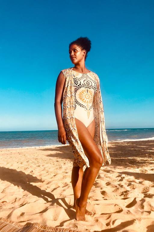 Uma mulher está posando na praia, vestindo um maiô branco e uma capa com estampas coloridas. Ela está de pé na areia, com o mar ao fundo e um céu azul claro. A luz do sol ilumina a cena, criando um ambiente tropical.
