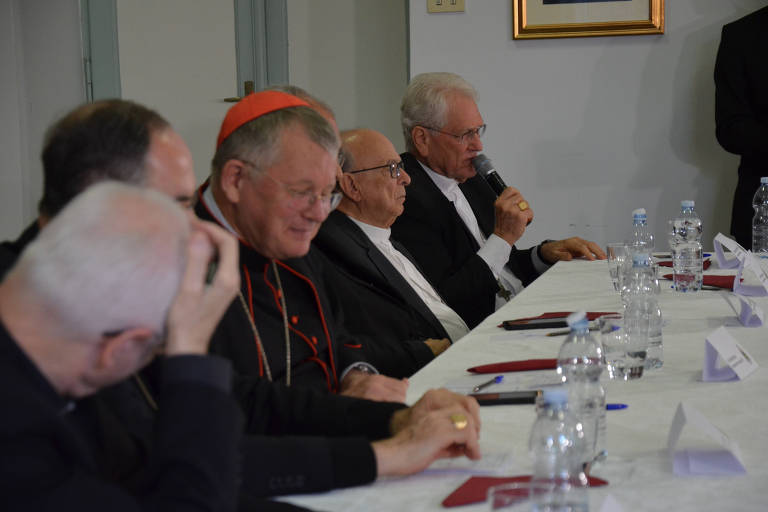 The image shows a table with several religious leaders seated. One of the men, identified as a cardinal, is wearing a red cassock and is on the left. Other men are seated around the table, some with serious expressions, while one of them speaks into a microphone. There are bottles of water and papers on the table, and in the background, a light wall with a framed painting.