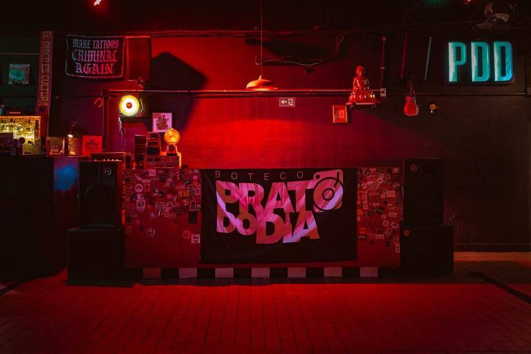 A imagem mostra o interior de um bar com iluminação em tons de vermelho e azul. No fundo, há uma parede decorada com várias fotos e adesivos. Um banner com o nome Pratododia está em destaque na parte frontal. Há também caixas de som e uma prateleira com objetos decorativos. O ambiente parece ser um local de entretenimento, possivelmente para música ao vivo.