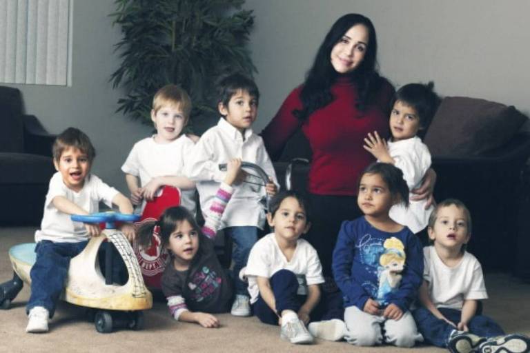 A imagem mostra um grupo de oito crianças sentadas no chão, vestindo camisetas brancas, com um adulto ao fundo. A mulher está usando uma blusa vermelha e está sorrindo. As crianças estão em várias poses, algumas sorrindo e outras olhando para a câmera. Ao lado de uma das crianças, há um brinquedo em forma de dinossauro. O ambiente é interno, com uma planta ao fundo e uma parede clara.