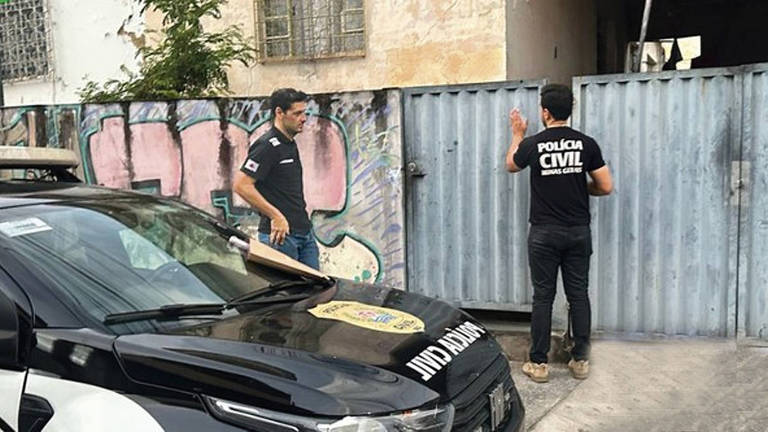A imagem mostra dois homens, um deles em uma viatura da Polícia Civil, enquanto o outro está em frente a um portão de metal. O homem na viatura parece estar conversando com o outro, que está de costas e usa uma camiseta preta com a inscrição 'POLÍCIA CIVIL'. Ao fundo, há uma parede com grafites coloridos.