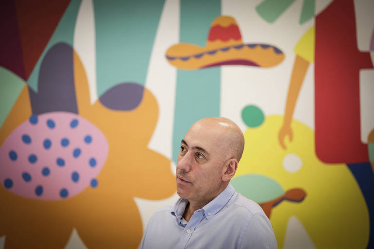 A imagem mostra um homem de cabeça careca, usando uma camisa azul clara, olhando para o lado. Ao fundo, há um mural colorido com formas abstratas, incluindo flores, um chapéu de sombrero e outras figuras em várias cores vibrantes.