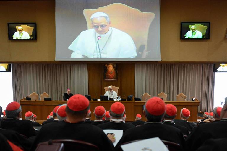A imagem mostra uma sala de conferência onde o Papa está falando. Ele está em um palco, com um microfone, e sua imagem é projetada em um telão atrás dele. Na plateia, há vários cardeais, todos usando vestes e chapéus vermelhos. A sala tem uma decoração simples, com cortinas e uma pintura na parede ao fundo.