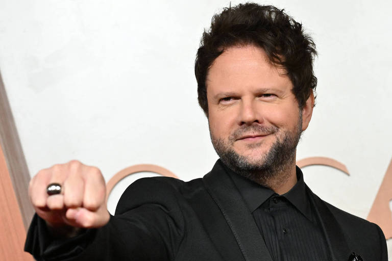 Um homem com cabelo cacheado e barba, vestindo um terno preto, está posando em um evento formal. Ele está fazendo um gesto com a mão, com o punho cerrado e o dedo indicador estendido. O fundo é neutro, com elementos que sugerem um evento de premiação.