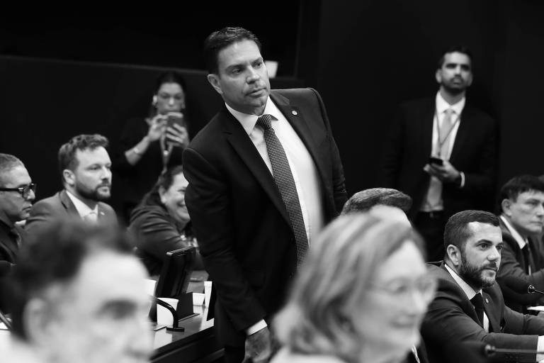 A imagem mostra uma reunião no Congresso Nacional do Brasil, com vários deputados e assessores presentes. Um homem em pé, vestido com um terno escuro e gravata, está se destacando no centro da imagem. Ao fundo, é possível ver outras pessoas sentadas, algumas olhando para o homem em pé. O ambiente é formal, com mesas e cadeiras dispostas em um formato que sugere uma sessão de trabalho.