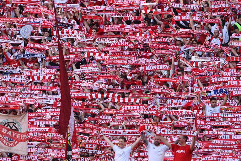 A imagem mostra uma grande quantidade de torcedores do Bayern de Munique em um estádio, segurando cachecóis vermelhos e brancos. Os cachecóis têm inscrições em letras brancas, e a maioria dos torcedores está vestida com camisetas brancas ou vermelhas. O ambiente é vibrante e cheio de energia, com muitos torcedores levantando os braços em celebração.