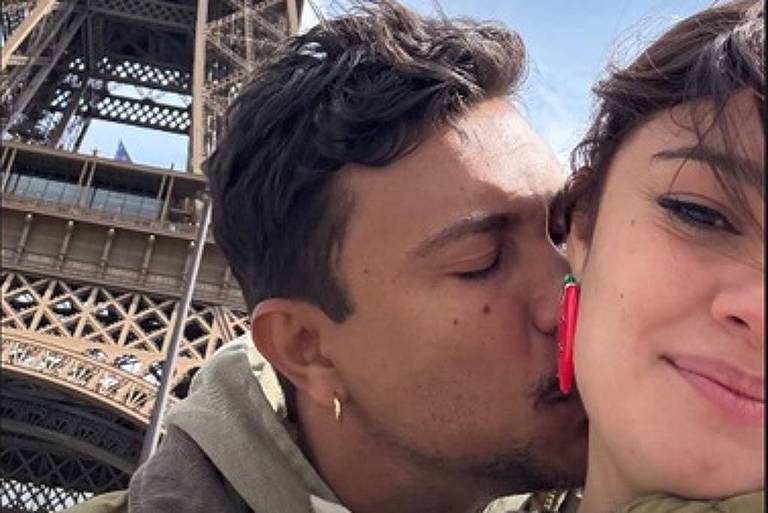 A imagem mostra um casal posando em frente à Torre Eiffel. O homem está dando um beijo na bochecha da mulher, que sorri para a câmera. O fundo é dominado pela estrutura da Torre Eiffel, com um céu parcialmente nublado.