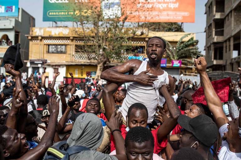 A imagem mostra uma multidão em um protesto, com várias pessoas levantando os braços. Um homem em destaque, sem camisa, é carregado por outros manifestantes. O ambiente é urbano, com prédios ao fundo e um grande cartaz visível. A atmosfera parece ser de agitação e energia coletiva.