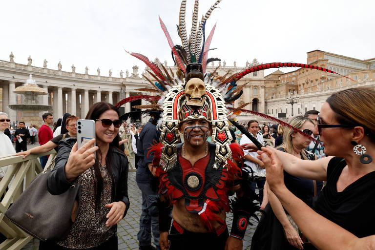 A imagem mostra um grupo de pessoas em uma praça, com destaque para um homem vestido com um traje tradicional colorido e um elaborado cocar de penas. Ele está posando para fotos com duas mulheres, uma delas segurando um celular. Ao fundo, é possível ver uma arquitetura clássica e várias outras pessoas observando a cena.
