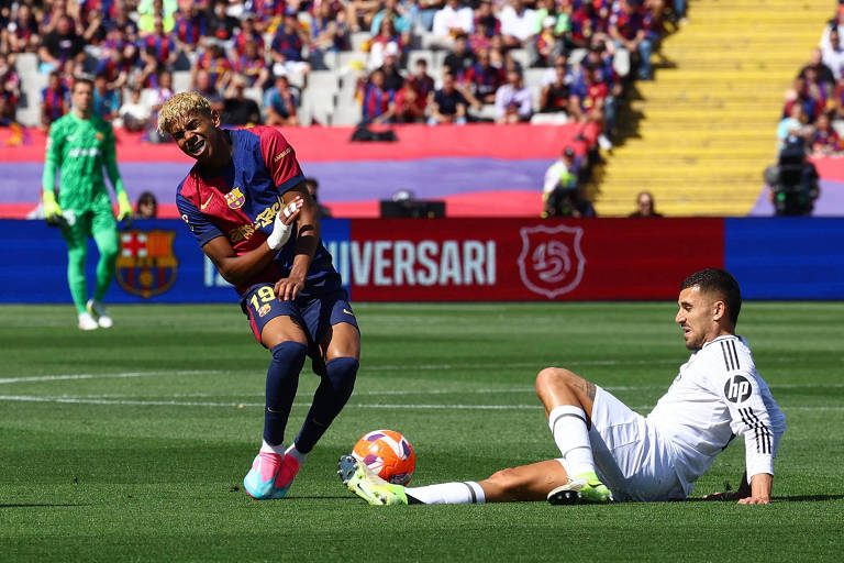 A imagem mostra um momento de um jogo de futebol entre o Barcelona e o Real Madrid. Um jogador do Barcelona, com cabelo cacheado e uniforme listrado em azul e grená, está em uma posição de queda, enquanto um jogador do Real Madrid, vestido de branco, está sentado no chão. Ao fundo, há uma multidão de torcedores e banners comemorativos. Um goleiro do Barcelona pode ser visto à esquerda, usando um uniforme verde.