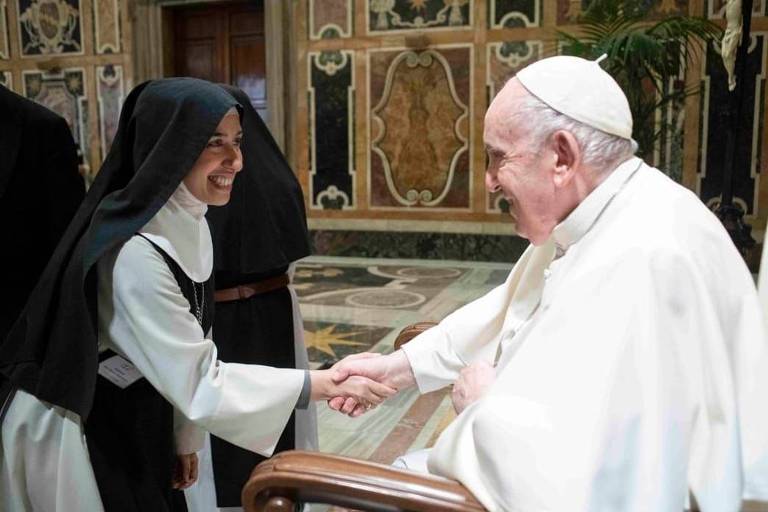 A imagem mostra o Papa Francisco sorrindo e apertando a mão de uma freira. A freira está vestida com um hábito tradicional e sorri enquanto cumprimenta o Papa. O fundo apresenta uma decoração elaborada, típica de um ambiente religioso, com detalhes em mármore e arte. O Papa está sentado em uma cadeira, enquanto a freira se inclina levemente para frente.