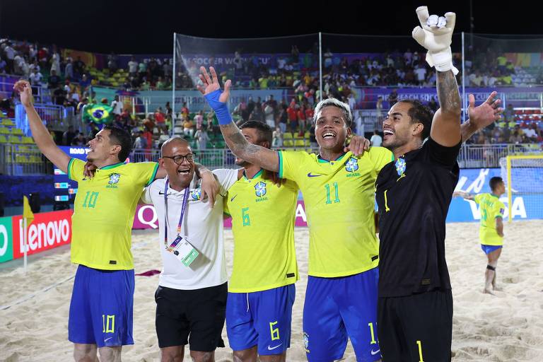 Cinco jogadores de futebol de areia da seleção brasileira estão em um campo de areia, celebrando uma vitória. Eles estão vestidos com uniformes amarelos e azuis, sorrindo e levantando os braços em sinal de alegria. Um dos jogadores é o goleiro, que está usando luvas e uma camisa preta. Ao fundo, há uma multidão assistindo ao jogo, com algumas bandeiras e banners visíveis.