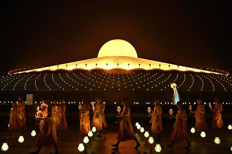 A imagem mostra um grupo de monges budistas vestidos com túnicas laranjas, caminhando em direção a uma grande estrutura iluminada que se assemelha a um templo ou monumento. A estrutura tem um grande domo amarelo no topo e está cercada por luzes brilhantes. Os monges seguram lanternas enquanto caminham em um padrão organizado, criando uma atmosfera serena e espiritual. O cenário é noturno, com um céu escuro ao fundo.