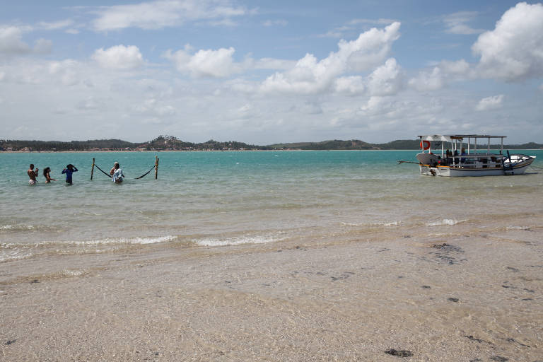A imagem mostra uma praia com areia clara e água azul. Algumas pessoas estão na água, aparentemente pescando ou realizando alguma atividade. Ao fundo, há um barco ancorado próximo à costa. O céu está parcialmente nublado, com algumas nuvens brancas.