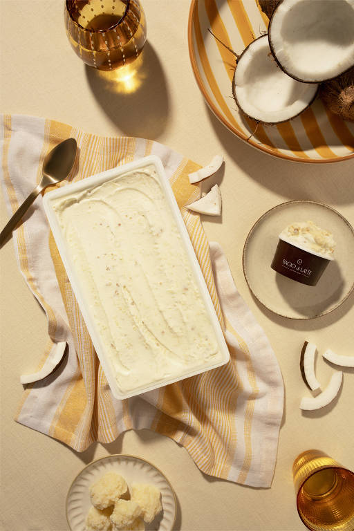 A imagem mostra um recipiente retangular de sorvete de coco, com uma colher ao lado, sobre um pano listrado em amarelo e branco. Ao redor, há pedaços de coco fresco, um prato com sorvete em uma tigela e um copo de bebida dourada. O fundo é claro e neutro, destacando os elementos da composição.
