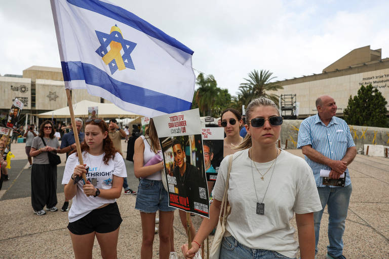 Um grupo de pessoas participa de um protesto ao ar livre. Algumas pessoas seguram cartazes, enquanto uma jovem à frente segura uma bandeira de Israel. O ambiente parece ser urbano, com edifícios ao fundo e palmeiras visíveis. As pessoas estão vestidas de maneira casual e algumas usam óculos de sol.